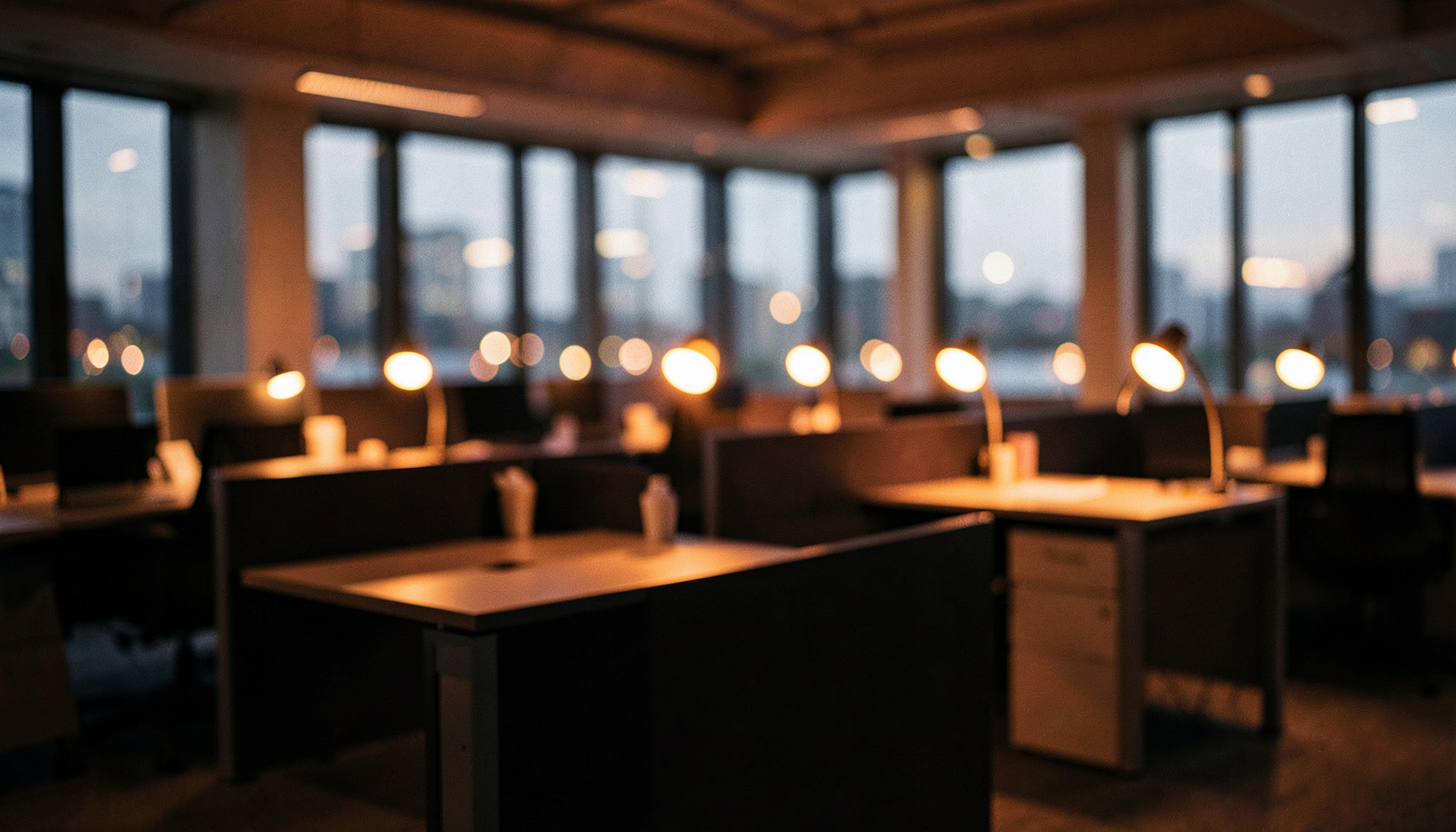 Wide cinematic frame of a modern open-plan office at dusk, warm amber desk lamps glowing against deep charcoal workstations, calm and focused mood